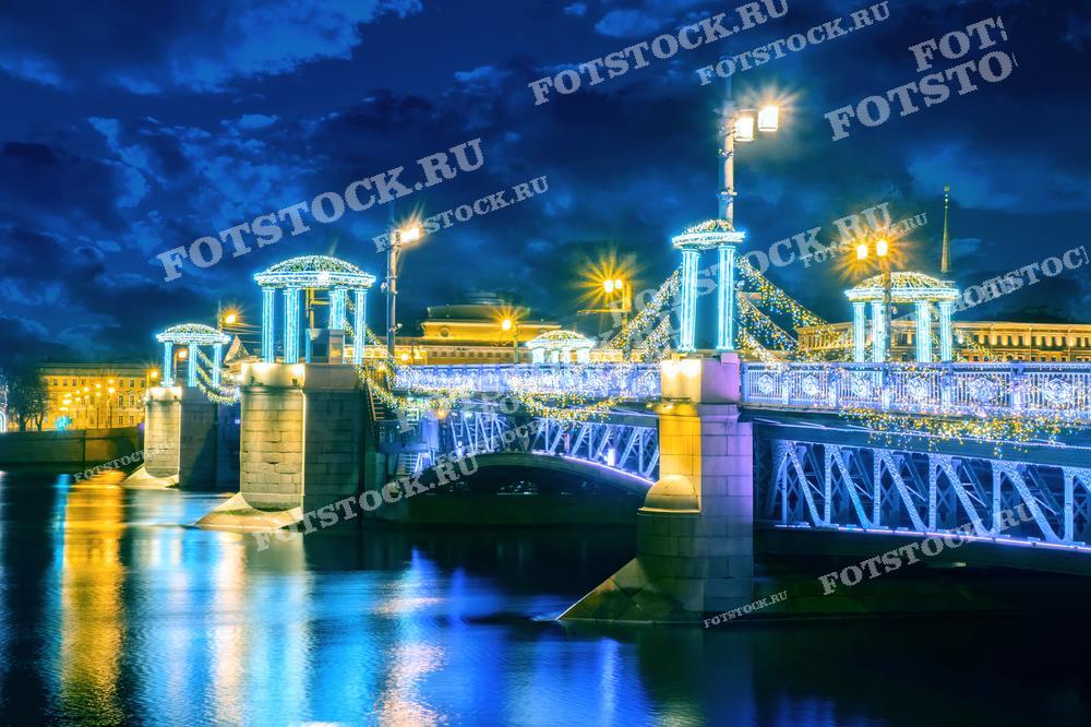 На фотографии изображен один из мостов Санкт-Петербурга ночью, украшенный яркой праздничной иллюминацией. Огни и гирлянды создают волшебную атмосферу, а их отражение на водной глади придает снимку дополнительную глубину и красоту. Этот вид подчеркивает праздничное настроение зимнего Петербурга.